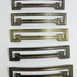 May include: Five antique drawer pulls in various metallic finishes, including bronze and brass. Each pull features a rectangular design with a decorative corner detail. The pulls are arranged vertically against a white background, showcasing their different patinas.