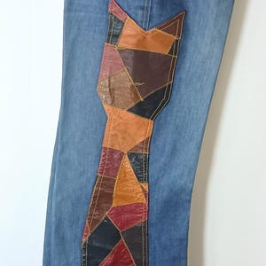 May include: Blue denim jeans featuring a large, vertical patchwork panel made of multi-colored leather segments in shades of brown, tan, red, and black. The leather patches are stitched onto the side of the leg and the waistband area.
