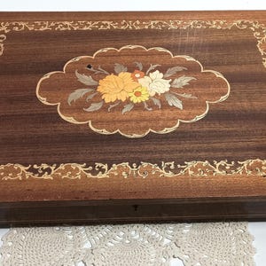 May include: A rectangular wooden jewelry box with a dark brown finish. The lid features a floral inlay with yellow and white flowers. Intricate light-colored scrollwork borders the lid and the floral design. A small keyhole is visible.