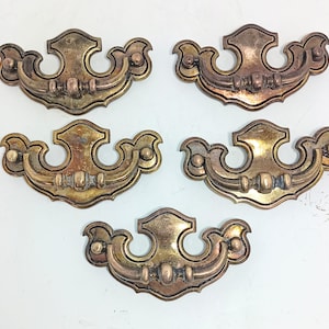 Puede incluir: Cinco tiradores de cajón antiguos de latón con un diseño ornamentado. Cada tirador presenta una forma de escudo central con asas curvas y detalles decorativos. Los tiradores tienen una pátina cálida y envejecida, lo que sugiere un uso vintage o reutilizado.