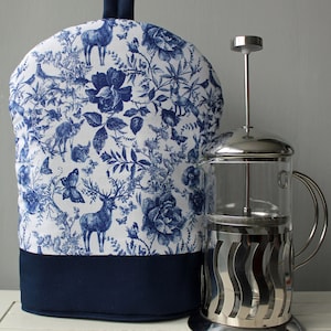May include: A blue and white floral patterned teapot cosy with a navy blue trim. The cosy is sitting on a white surface next to a silver French press.