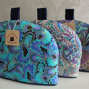 Peut inclure: Trois chauffe-thé en tissu à motifs bleus, violets et dorés avec des poignées bleu marine. L'étiquette sur le chauffe-thé le plus à gauche indique "Made with Love by Crafty Dora UK".