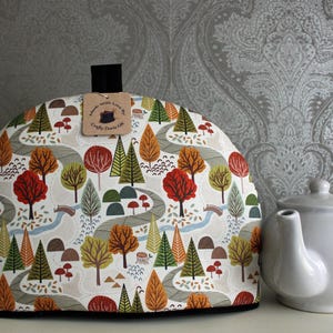 May include: A white tea cozy with a woodland pattern featuring trees, leaves, and a winding path. The cozy has a black trim and a brown tag that reads "Handmade with Love by Crafty Diva UK".