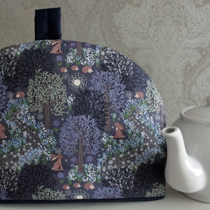 May include: A blue and white teapot cozy with a woodland pattern featuring trees, flowers, and rabbits. The cozy is made of fabric and has a dark blue trim.