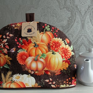 May include: A decorative tea cosy featuring a pumpkin and floral design in autumnal colours. A white teapot sits beside the cosy. The cosy has a tag that says "Made with Love by Crafty Doris UK."