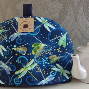 Peut inclure: Un chauffe-thé en tissu bleu avec un motif de libellules et des accents de paillettes dorées. Le chauffe-thé a une poignée bleu marine et une étiquette brune qui indique "Handmade with Love by Crafty Doris UK".