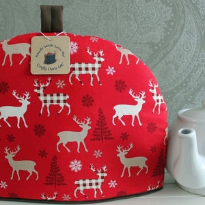 May include: A festive red tea cosy featuring a pattern of white deer, snowflakes, and trees. It has a brown handle and trim. A white teapot sits beside it. The tag reads "Made with Love by Crafty Doris UK".