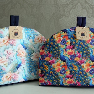 May include: Two tea cosy covers featuring a peacock and floral design. The covers are blue, green and pink with a navy blue trim. The covers have a tag that says "Handmade with Love, Crafty Dawn Life".