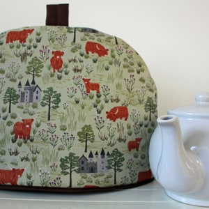 May include: A green and brown tea cosy with a pattern of red Highland cows and castles. The cosy is on a white surface next to a white teapot.