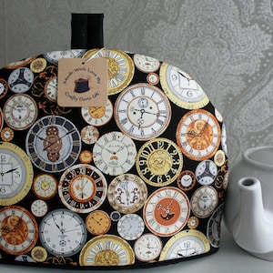 May include: A black tea cozy with a design of various clocks in gold, white, and black on a black background. The text "Made With Love By Crafty Doris UK" is on a white tag attached to the cozy.