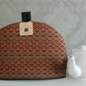 Op de afbeelding: Een theemuts met patroon met een zwart handvat en een witte theepot. De theemuts heeft een herhalend geometrisch ontwerp in rood, groen en goud. Een klein label is aan de theemuts bevestigd.