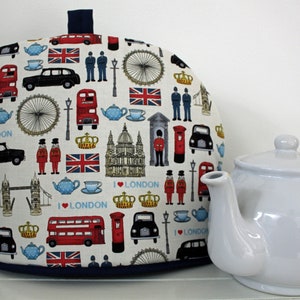 May include: A white teapot with a cream-coloured tea cosy featuring a London-themed print. The print includes red double-decker buses, black taxis, the Union Jack flag, and the words "I Love London".