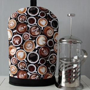 May include: A black and brown fabric coffee pot cover with a pattern of coffee cups and coffee beans. The cover is sitting on a white surface next to a silver French press.