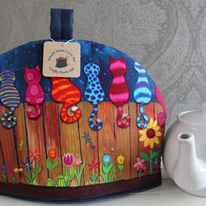 May include: A colourful tea cosy featuring a design of cats sitting on a fence. The cats are various colours, including green, blue, pink, orange, and purple. The background is a dark blue with stars. The text on the tag reads "Made with Love by Crafty Doris UK".