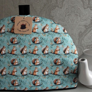 May include: A teal blue tea cozy with a pattern of squirrels. The cozy has a black handle and a brown tag that reads "Made with Love by Crafty Doris UK".