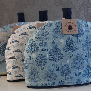 Peut inclure: Trois chauffe-thé en tissu bleu et blanc avec des motifs différents. Les housses sont en tissu avec un motif de forêt bleu et blanc. L'étiquette sur la housse de droite indique "Made with Love by Crafty Dears UK".