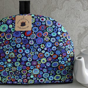May include: A colorful tea cozy with a black background and a pattern of blue, green, orange, and purple circles. The tea cozy has a black handle and a tag that reads "Handmade With Love By Crafty Doris UK".