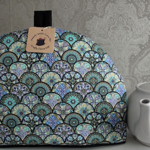 May include: A teal, blue, and gold patterned tea cozy with a black handle and a brown tag that reads "Made with Love by Crafty Doris UK".