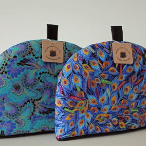 Könnte beinhalten: Zwei halbkreisförmige Teewärmer aus Stoff mit lebhaften Mustern. Der linke Teewärmer hat ein blaugrünes und violettes Paisley-Design, während der rechte ein blau-oranges Pfauenfeder-Motiv aufweist. Beide haben eine schwarze Schlaufe und ein Etikett.