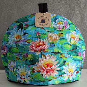 May include: A teal blue tea cozy with a floral pattern of water lilies. The cozy has a black handle and a brown tag that reads "Made with Love by Crafty Doris UK".