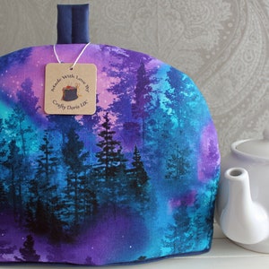 May include: A purple and blue tea cosy with a forest and starry sky design. The cosy has a navy blue handle and a tag that reads "Made with Love by Crafty Doris UK".