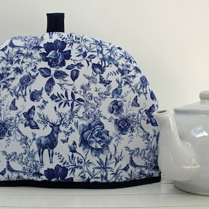May include: A white teapot with a blue and white floral patterned tea cosy. The tea cosy has a blue deer and butterfly design.