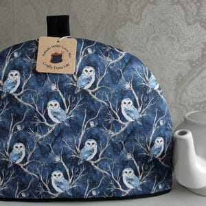May include: A blue tea cozy with a white owl pattern. The cozy has a black trim and a tag that reads "Made with Love by Crafty Doris UK".