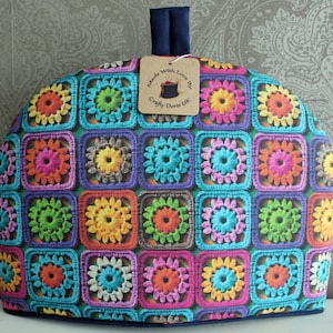 Op de afbeelding: Een kleurrijke gehaakte theekos met een vierkant patroon. De theekos heeft een blauwe handgreep en een label met de tekst "Made with Love by Crafty Doris UK".