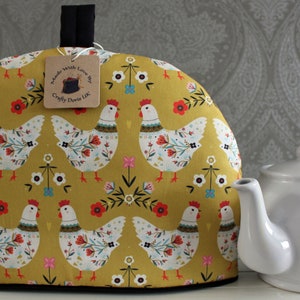 May include: A yellow tea cosy with a pattern of colourful chickens and flowers. The text "Made with Love By Crafty Owls UK" is on a tag attached to the cosy.
