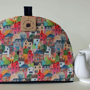 Peut inclure: Une housse de théière en forme de dôme avec un motif coloré de maisons et d'arbres dans des tons de rouge, bleu et vert. Une boucle en tissu bleu foncé se trouve au sommet avec une étiquette en carton brun indiquant Made With Love By Crafty Doris LLC.