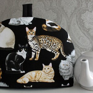 May include: Black tea cosy with a colourful cat print. The cosy features a variety of cats in different poses, including a tabby cat, a Siamese cat, and a black and white cat. The cosy has a handle on the top.