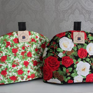 Op de afbeelding: Twee theemutsen met bloempatronen. Eén heeft een witte achtergrond met rode rozen, groene bladeren en dennenappels. De andere heeft een zwarte achtergrond met rode en witte rozen, groene bladeren en dennenappels. Elk heeft een zwart handvat en een label.