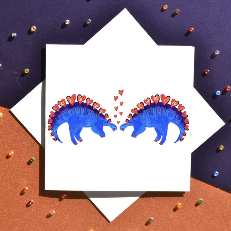 K&ouml;nnte beinhalten: Eine wei&szlig;e Karte mit zwei blauen Dinosauriern, die sich gegen&uuml;berstehen. Die Dinosaurier haben rote Herzen auf ihren R&uuml;cken, die die Namen "Clair" und "Chris" buchstabieren.