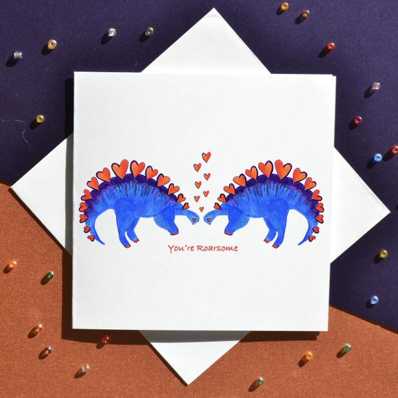 K&ouml;nnte beinhalten: Eine Gru&szlig;karte mit zwei blauen Dinosauriern mit roten Herzen auf ihren R&uuml;cken, die sich gegen&uuml;berstehen. Die Dinosaurier k&uuml;ssen sich und Herzen schweben &uuml;ber ihnen. Der Text auf der Karte lautet "You're Roarsome".