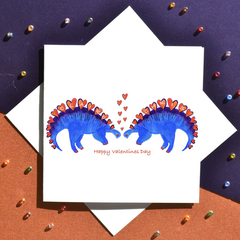 K&ouml;nnte beinhalten: Eine Valentinstagskarte mit zwei blauen Dinosauriern, deren rote Herzen die Namen "Claire" und "Chris" buchstabieren. Die Dinosaurier schauen sich an und haben ihre H&auml;lse so gebogen, als w&uuml;rden sie sich k&uuml;ssen. Der Text "Happy Valentines Day" ist unter den Dinosauriern gedruckt.