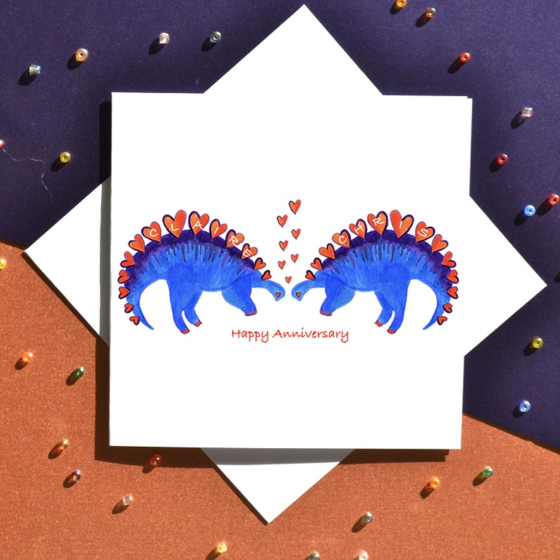 K&ouml;nnte beinhalten: Eine wei&szlig;e Karte mit zwei blauen Dinosauriern, die sich gegen&uuml;berstehen. Die Dinosaurier haben rote Herzen auf ihren R&uuml;cken, die die Namen "Claire" und "Chris" buchstabieren. &Uuml;ber den Dinosauriern befinden sich rote Herzen. Die Karte lautet "Happy Anniversary".