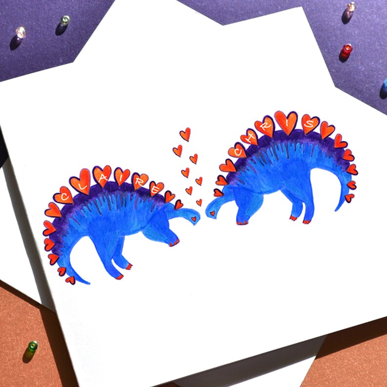K&ouml;nnte beinhalten: Zwei blaue Dinosaurier mit orangefarbenen Herzen auf ihren R&uuml;cken, die sich gegen&uuml;berstehen. Der Dinosaurier links sagt "CLAIRE" und der Dinosaurier rechts sagt "CHRIS". Zwischen den Dinosauriern schweben kleine rote Herzen.