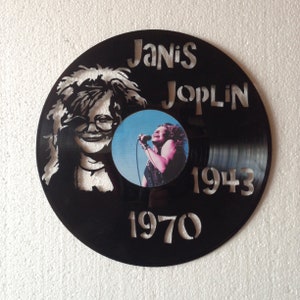 May include: A black vinyl record with a white silhouette of Janis Joplin's face and the text "Janis Joplin 1943 1970". A smaller photo of Janis Joplin singing is in the center of the record.