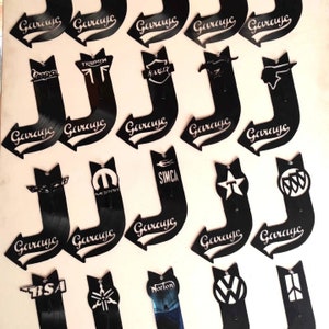 Könnte beinhalten: Zwanzig schwarze Vinyl-Schilder in Pfeilform mit weißem Text und Logos. Die Schilder zeigen "Garage" und verschiedene Auto- und Motorradlogos, darunter Triumph, Harley Davidson, Vespa und mehr.