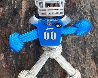 May include: A white paracord lanyard with a blue and silver Detroit Lions football player charm. The charm has the NFL logo, the word "Lions", and the number "00" on the jersey. The lanyard is tied in a knot at the bottom.