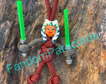 May include: A red paracord keychain with a Lego minifigure of Ahsoka Tano holding two green lightsabers. The keychain is tied in a knot and has a loop at the top for attaching to a bag or keys.