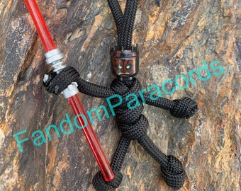 May include: A black paracord keychain figure holding a red lightsaber. The figure has a black head with a green and orange face. The keychain has a loop at the top for attaching to a bag or keys. The text "FandomParacords" is visible in the image.