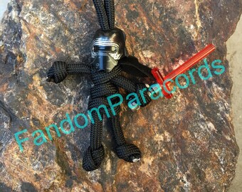 May include: Black paracord keychain with a Kylo Ren figure and a red lightsaber. The keychain is on a rock background with the text "FandomParacords".
