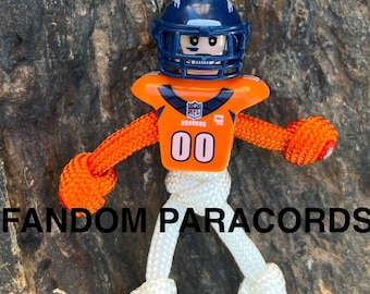 May include: A handcrafted Denver Broncos football player figurine made from orange and blue plastic and white paracord. The figure has a blue helmet and the number 00 on the jersey. The text "FANDOM PARACORDS" is visible.