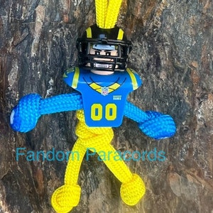 Puede incluir: Un llavero de paracord amarillo y azul con una figura de jugador de fútbol que lleva una camiseta azul y amarilla con el número "00" y la palabra "RAMS".