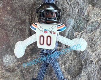 May include: A blue paracord keychain with a white football player figurine. The figurine is wearing a helmet, jersey, and pants. The jersey has the number "00" on the front and the word "Bears" on the chest.