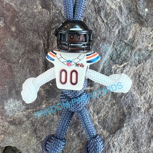 May include: A blue paracord keychain with a white football player figurine. The figurine is wearing a helmet, jersey, and pants. The jersey has the number "00" on the front and the word "Bears" on the chest.