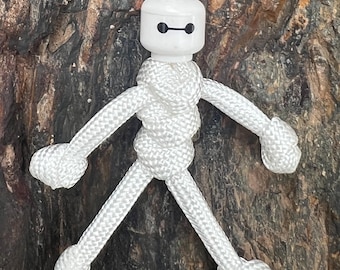 May include: A white rope keychain featuring a small, stylized figure with a white head and body. The figure has a simple design with arms and legs, and is suspended from a loop of white rope. The background is a textured, brown surface.