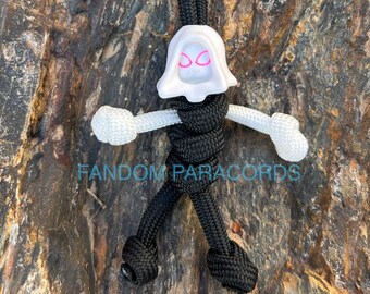May include: A white paracord figure with black paracord limbs and a white head with pink eyes. The figure is tied in a standing pose with arms outstretched. The figure is on a brown and grey rock surface. The text "FANDOM PARACORDS" is visible below the figure.