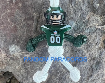 May include: A green and white paracord keychain with a New York Jets football player figurine. The figurine is wearing a helmet, jersey, and pants. The keychain is tied in a knot and has a loop at the top.